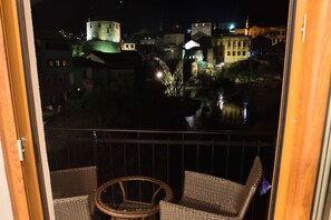 Un balcon agrémenté de fauteuils en osier, offrant une vue sur une ville dont les bâtiments sont illuminés, ainsi que sur une structure ressemblant à un château.