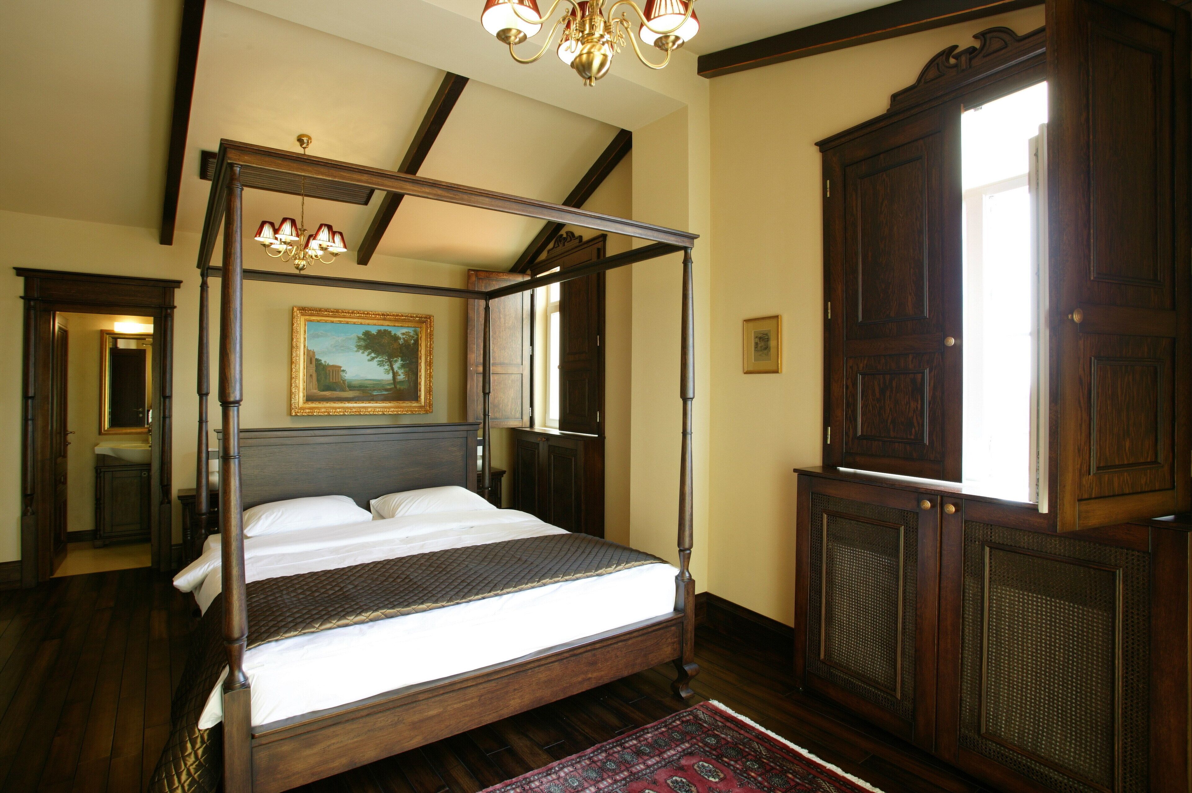 A four-poster bed with a dark wooden frame and white linens, a large mirror above the bed, and a painting on the wall.