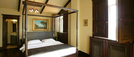 A four-poster bed with a dark wooden frame and white linens, a large mirror above the bed, and a painting on the wall.
