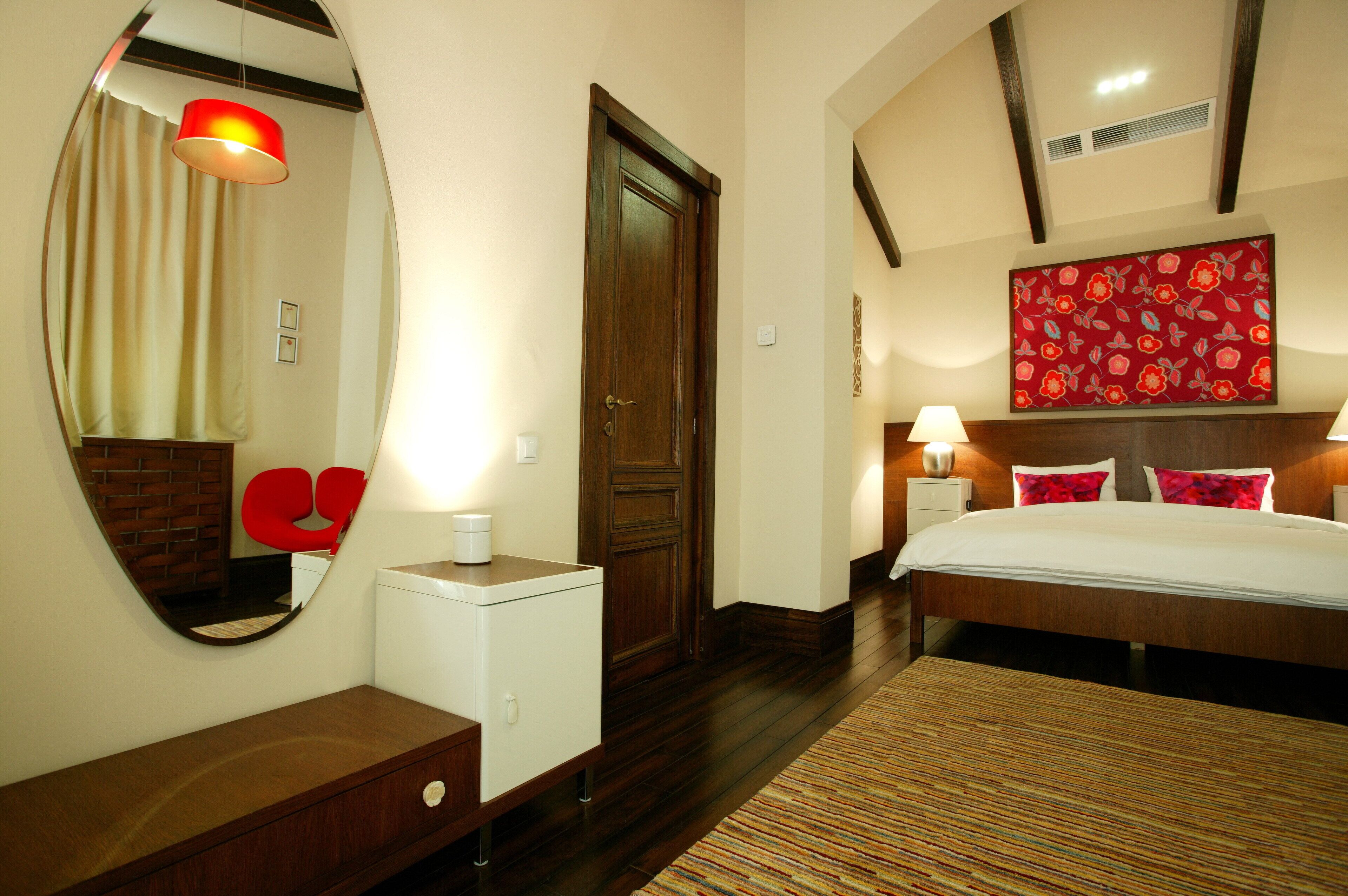A hotel room with a wooden floor, a bed with white bedding, a red floral painting on the wall, a mirror, and a red chair.