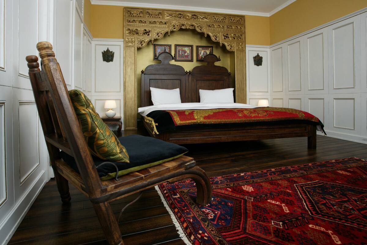 Une chambre à coucher de style traditionnel, avec un lit à baldaquin, une chaise en bois dotée d’un coussin et un tapis à motifs.