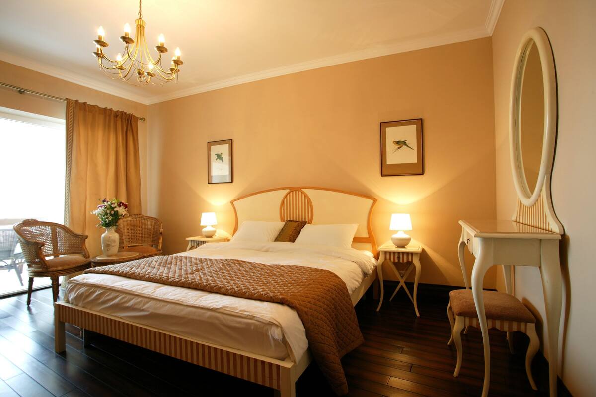 Une chambre bien éclairée, avec un grand lit, deux tables de chevet, une chaise, un miroir et une fenêtre avec des rideaux.