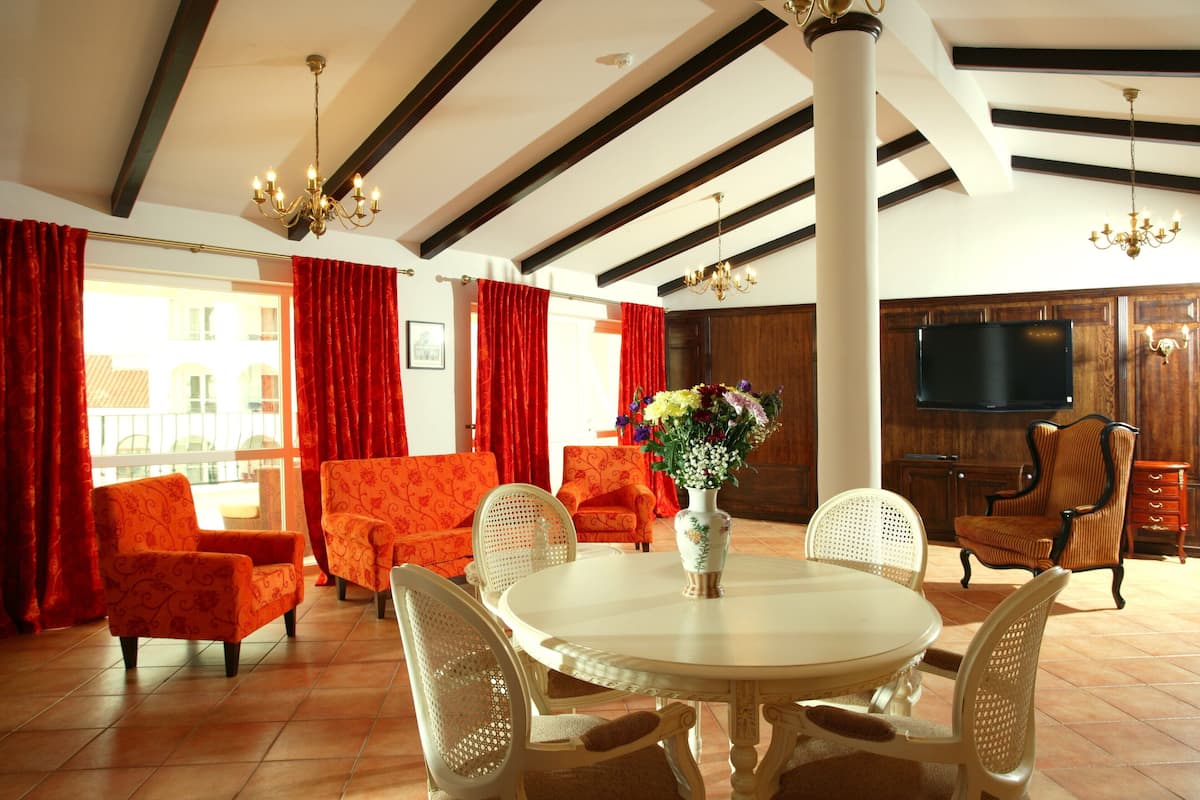 Un salon spacieux avec une table ronde, des canapés à motifs orange et une grande fenêtre ornée de rideaux rouges.
