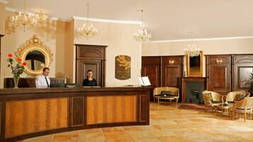 A reception area with a wooden reception desk, two chandeliers, a fireplace, and wicker furniture.