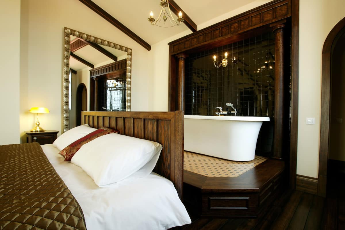 Une chambre d’hôtel avec un grand lit, une tête de lit en bois, un grand miroir, une baignoire et un lustre.