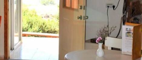 Una habitación con una mesa y sillas blancas, un pequeño florero blanco con flores, una puerta de madera que da a un área exterior y un televisor montado en la pared.