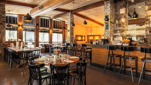A restaurant with high ceilings, exposed beams, and a bar area. Tables are set with chairs, and there are large windows allowing natural light.