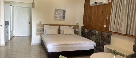A hotel room with a bed, a small table, a chair, a painting on the wall, and an air conditioning unit.
