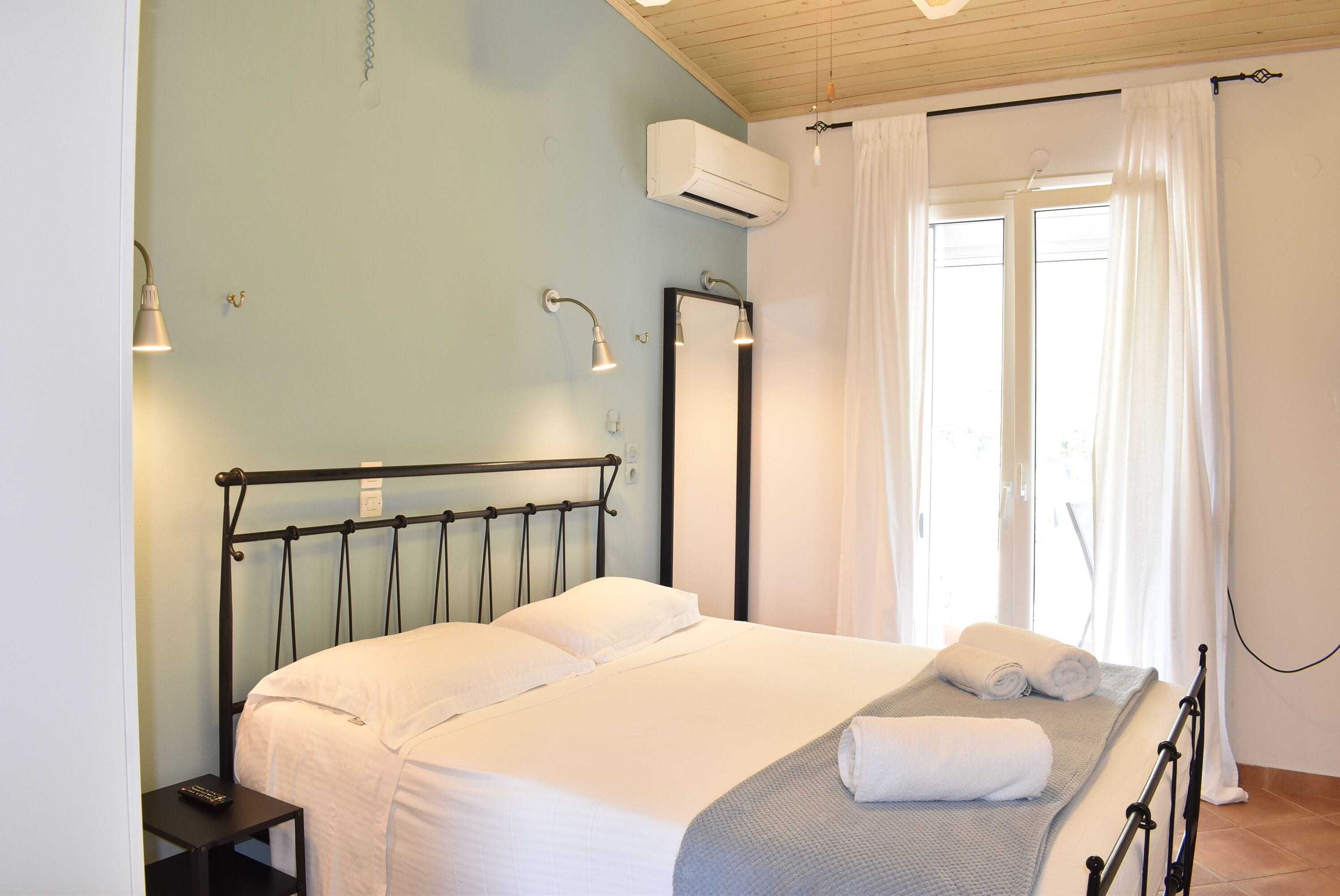 A neatly made bed with white linens, a bedside table with a remote control, and a window with white curtains.