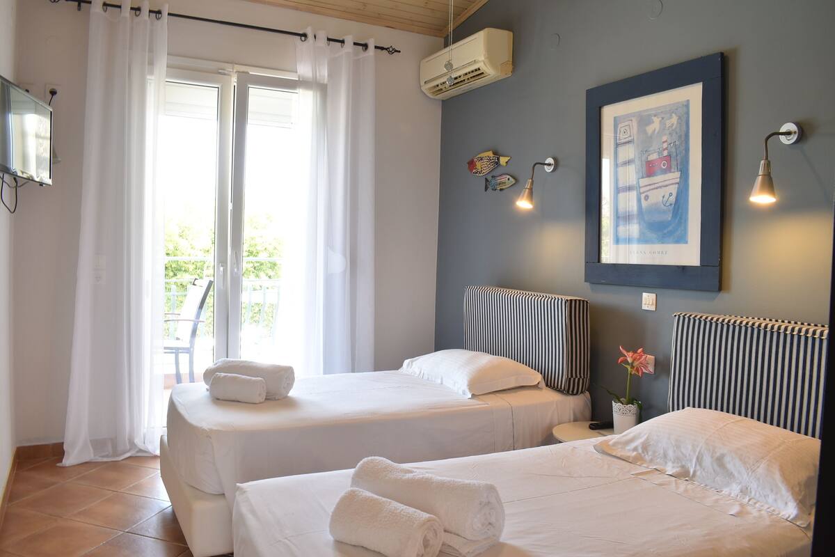 A hotel room with two beds, a large window with curtains, a wall-mounted air conditioner, a framed picture, and a small potted plant.