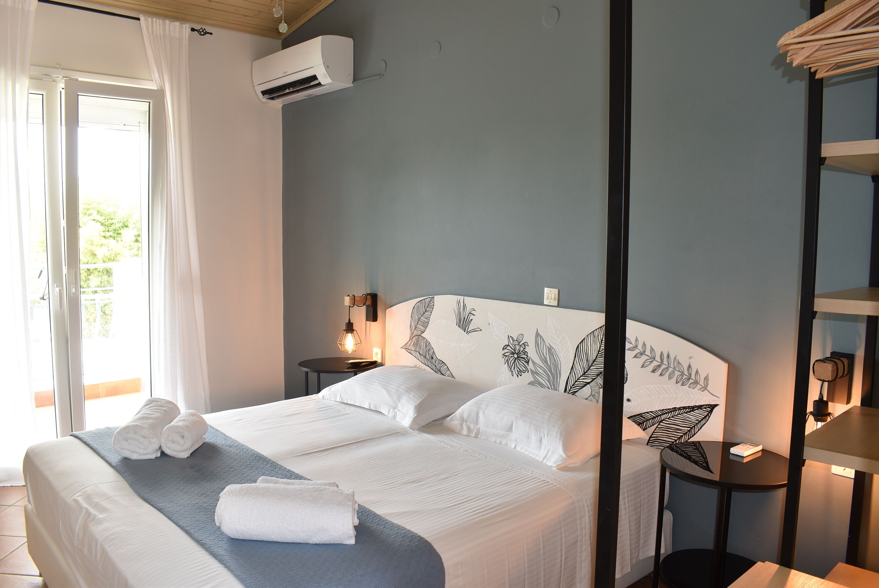A hotel room with a bed, a bedside table, an air conditioning unit, and a view of the outdoors.