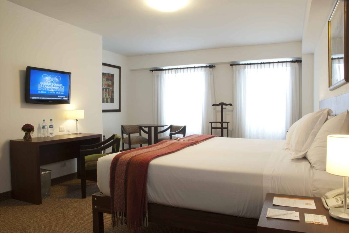 Una habitación de hotel con cama, escritorio, silla, televisor y cortinas.