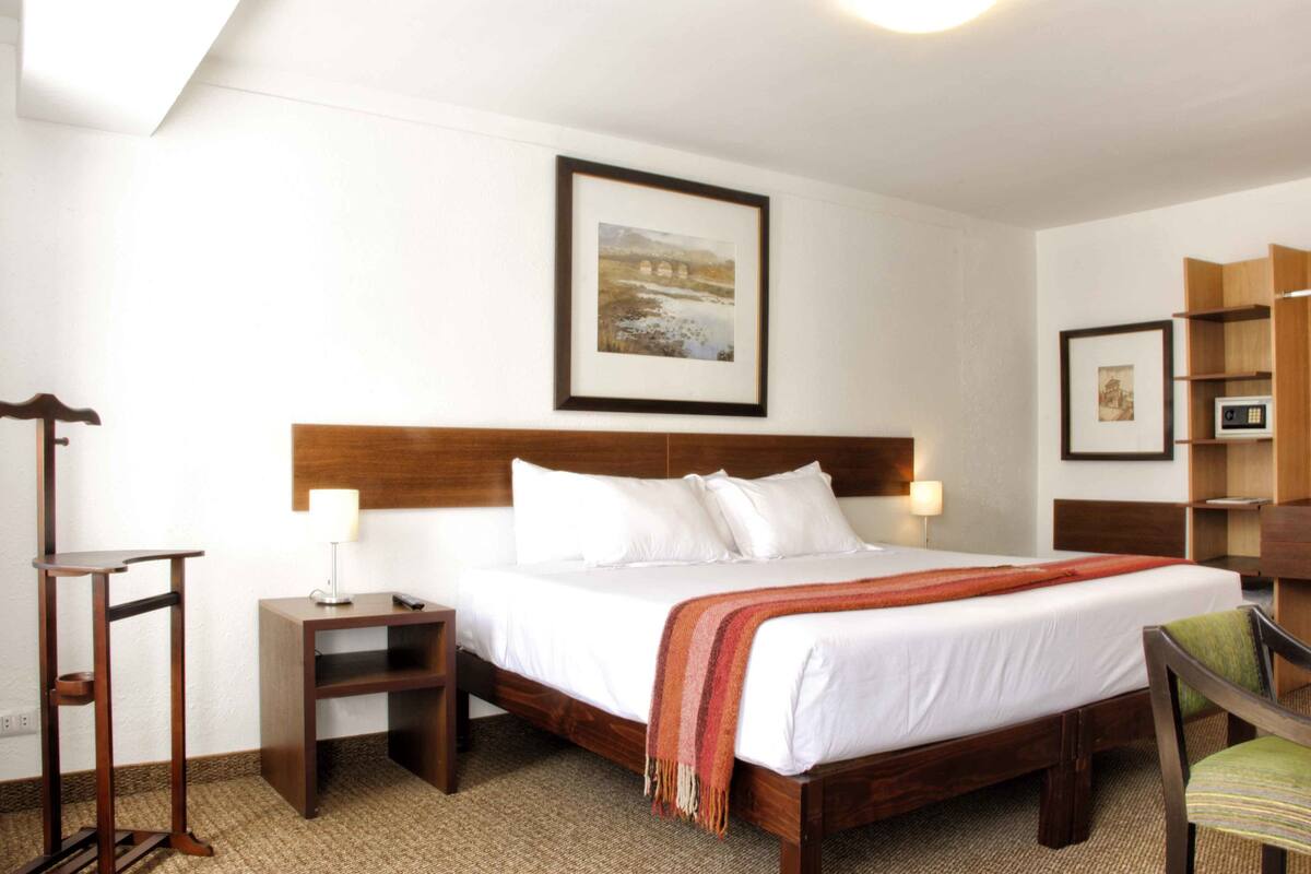 Habitación de hotel con cama de madera, ropa de cama blanca, un cuadro en la pared, una mesita de noche, una silla y un pequeño estante de madera.
