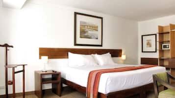 A hotel room with a wooden bed, white linens, a framed picture on the wall, a nightstand, a chair, and a small wooden shelf.