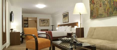 A hotel room with a bed, a sofa, a chair, a coffee table, a lamp, a painting, and a television.