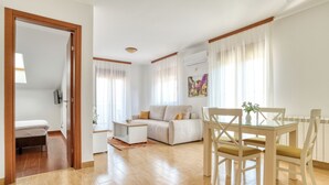 A modern living room with a sofa, a glass dining table, and chairs. There is a door leading to another room and an air conditioning unit on the wall. - Garni Hotel Milica (Denovici)