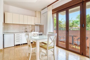 A modern kitchen with white cabinets, a glass dining table, and chairs, leading to an outdoor patio with a view of trees and a building.
