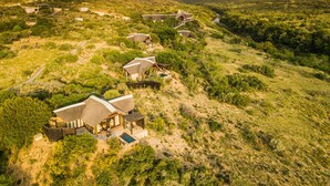 A series of thatched-roof cabins with outdoor seating areas, set in a hilly, green landscape with a river running through it. - Kwandwe Great Fish River Lodge (Makhanda)