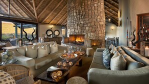 A cozy living room with a stone fireplace, comfortable seating, and a view of the outdoors. - Kwandwe Great Fish River Lodge (Makhanda)