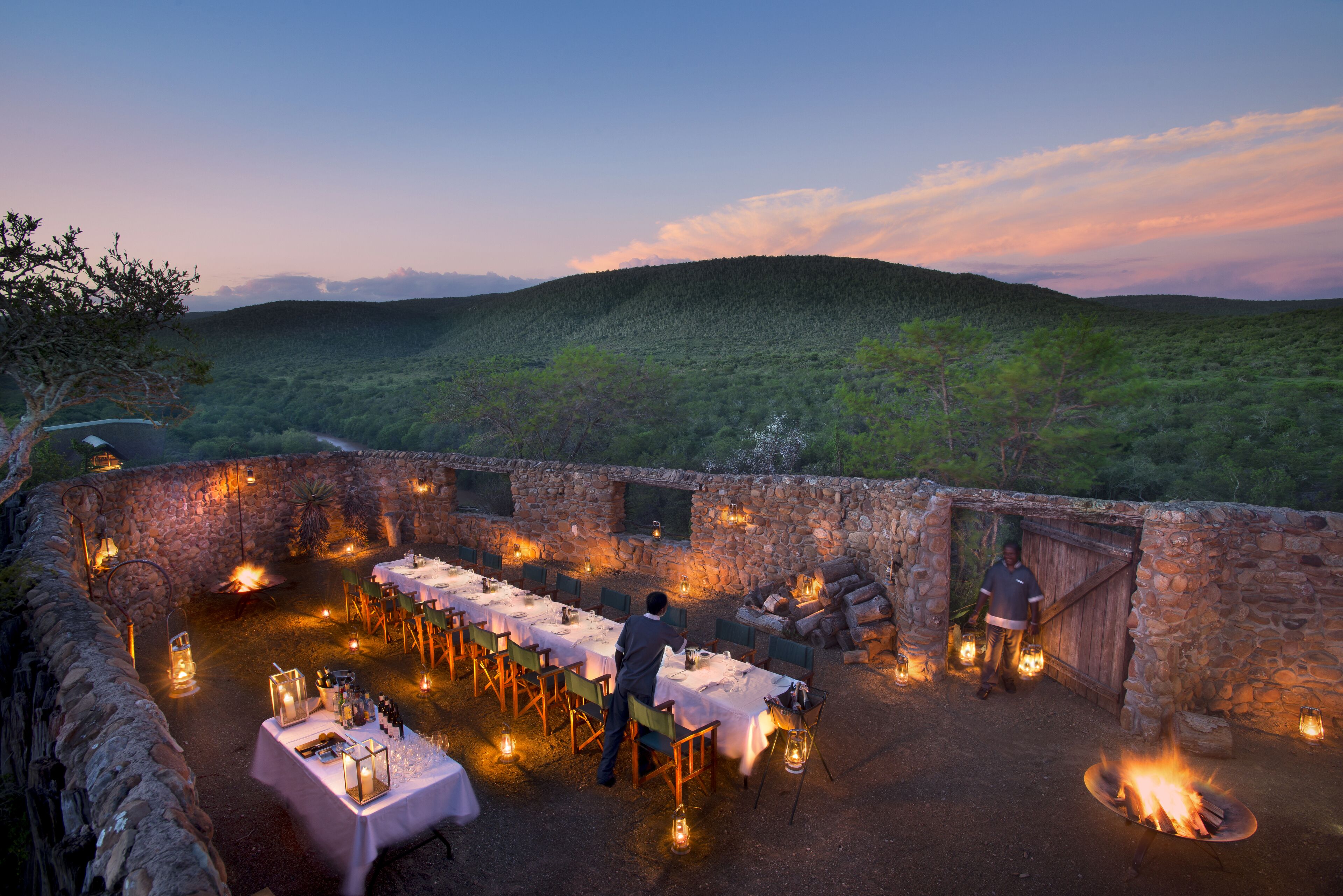 An outdoor dining area with tables set for a meal, surrounded by stone walls and lanterns, overlooking a forested landscape.