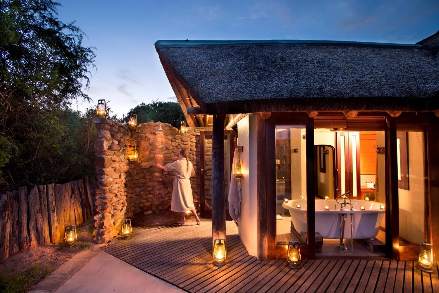 Une salle de bain extérieure aux murs de pierre, dotée d’une terrasse en bois, d’une baignoire et de lanternes.