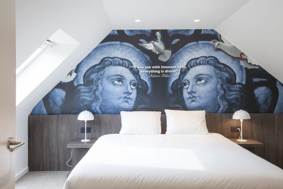 A bedroom with a large mural on the ceiling, a bed with white linens, two bedside tables with lamps, and a skylight.