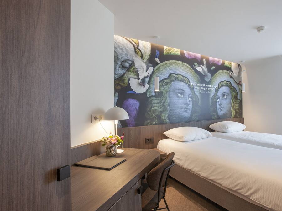 A hotel room with a large wall mural, a desk with a lamp, and a bed with white linens.