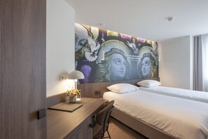 Ein Hotelzimmer mit einer großen Wandmalerei, einem Schreibtisch mit Lampe und einem Bett mit weißen Leinentüchern.