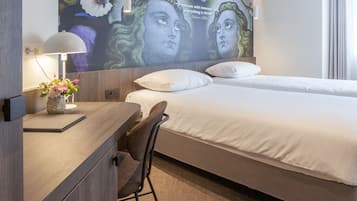 A modern hotel room with a large bed, a desk with a lamp, and a wall mural.