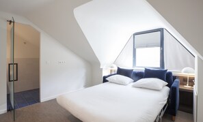 Ein Schlafzimmer mit schrägen Wänden, einem Bett mit weißer Bettwäsche, einem Schreibtisch mit Lampe und einem Fenster mit Jalousien.