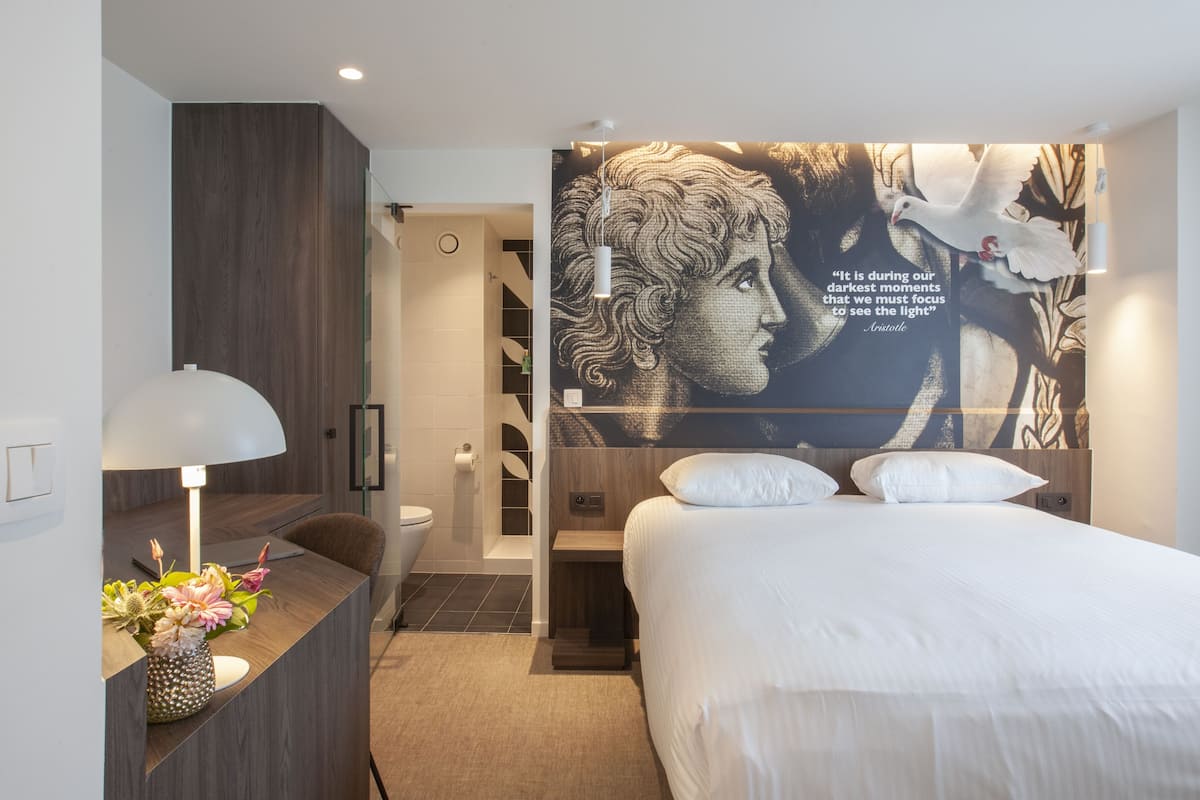 A modern hotel room with a bed, a desk with a lamp, and a wall with a large quote.