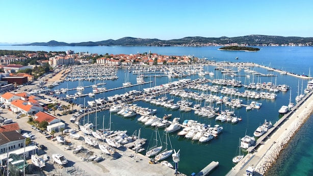 A marina with numerous boats docked, surrounded by buildings and a clear view of the sea and distant hills. - Apartments Moreta (Biograd na Moru)