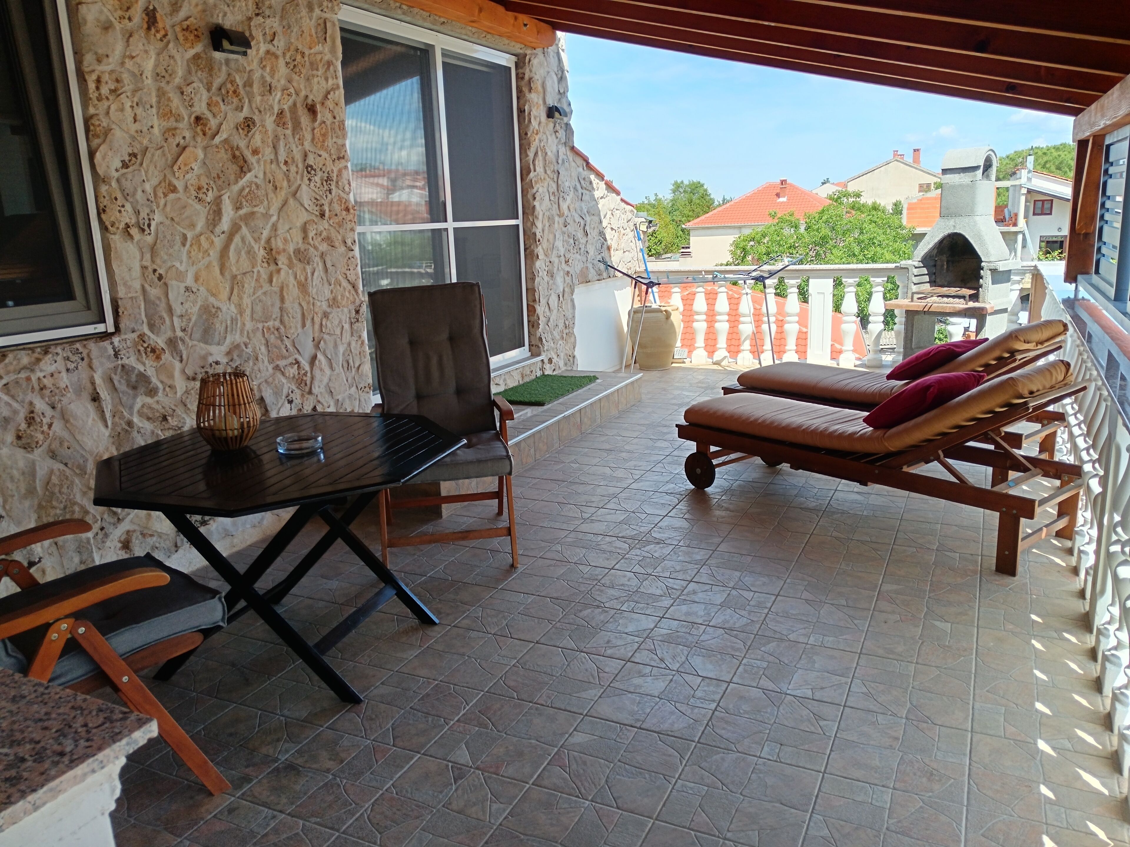A covered patio with a stone wall, a table with chairs, and two sun loungers with cushions.