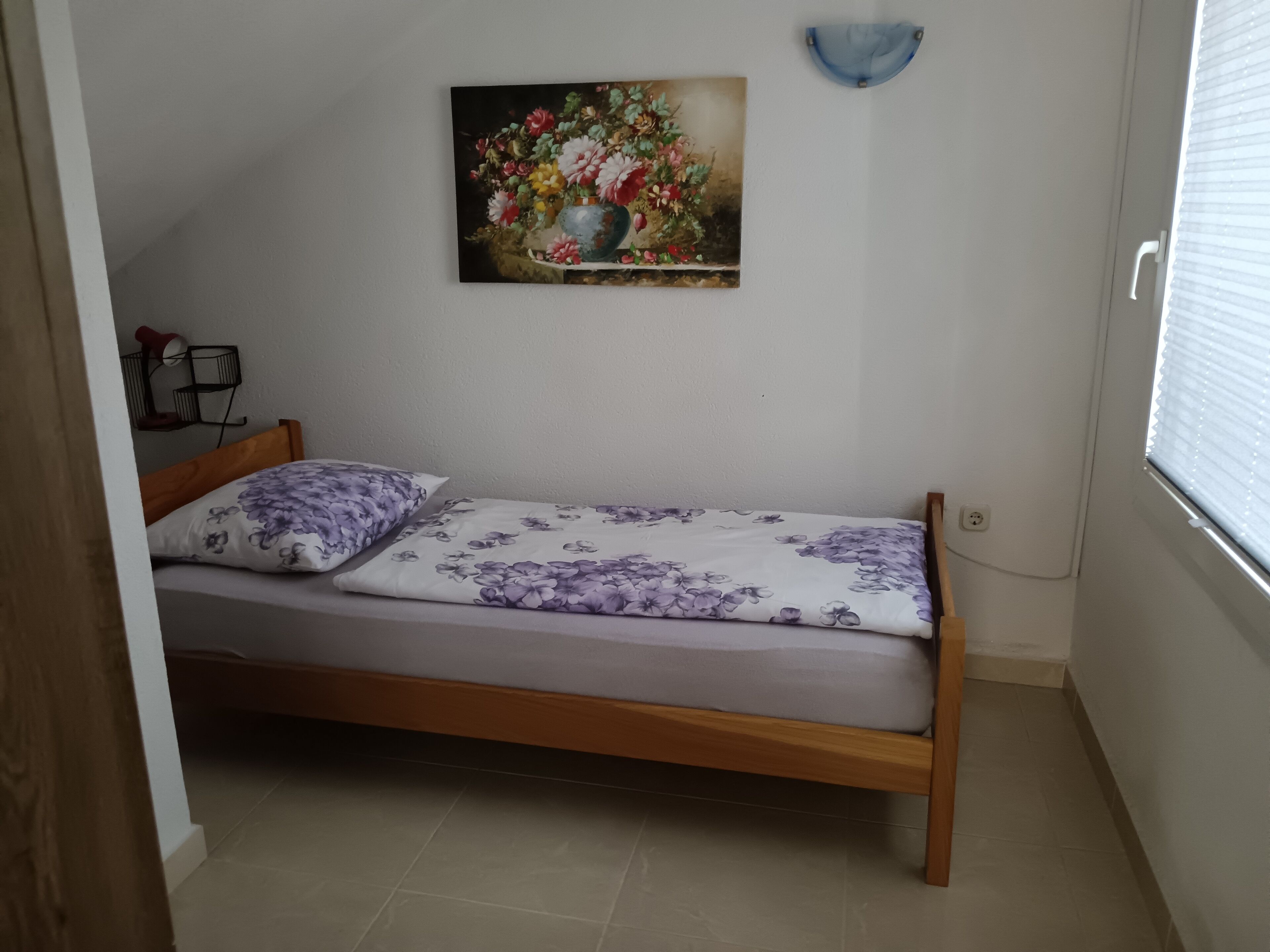 A small, single-bed room with a wooden frame, a floral-patterned bedspread, and a painting of flowers on the wall.