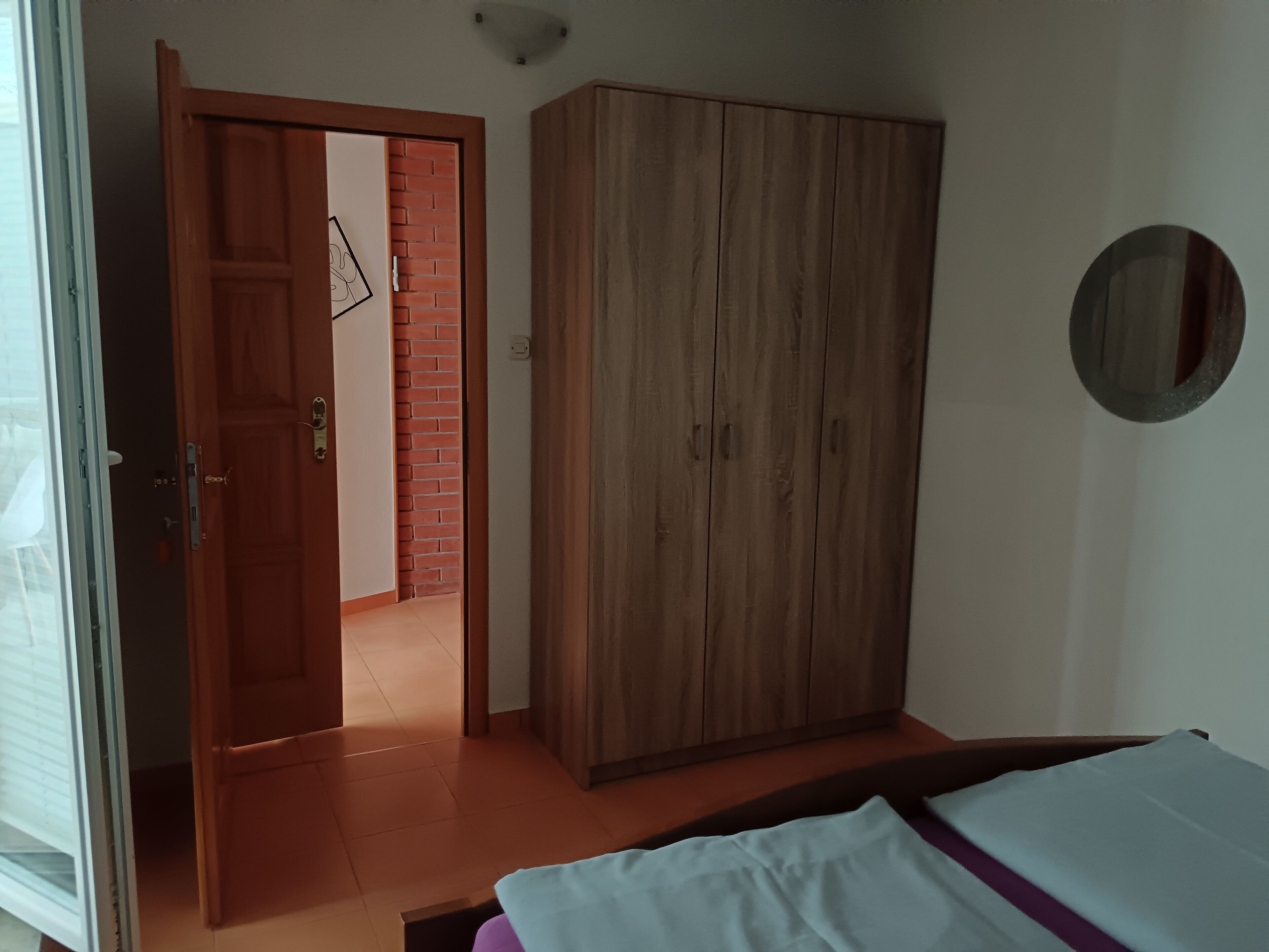 A bedroom with a wooden wardrobe, a bed, and a door leading to another room.