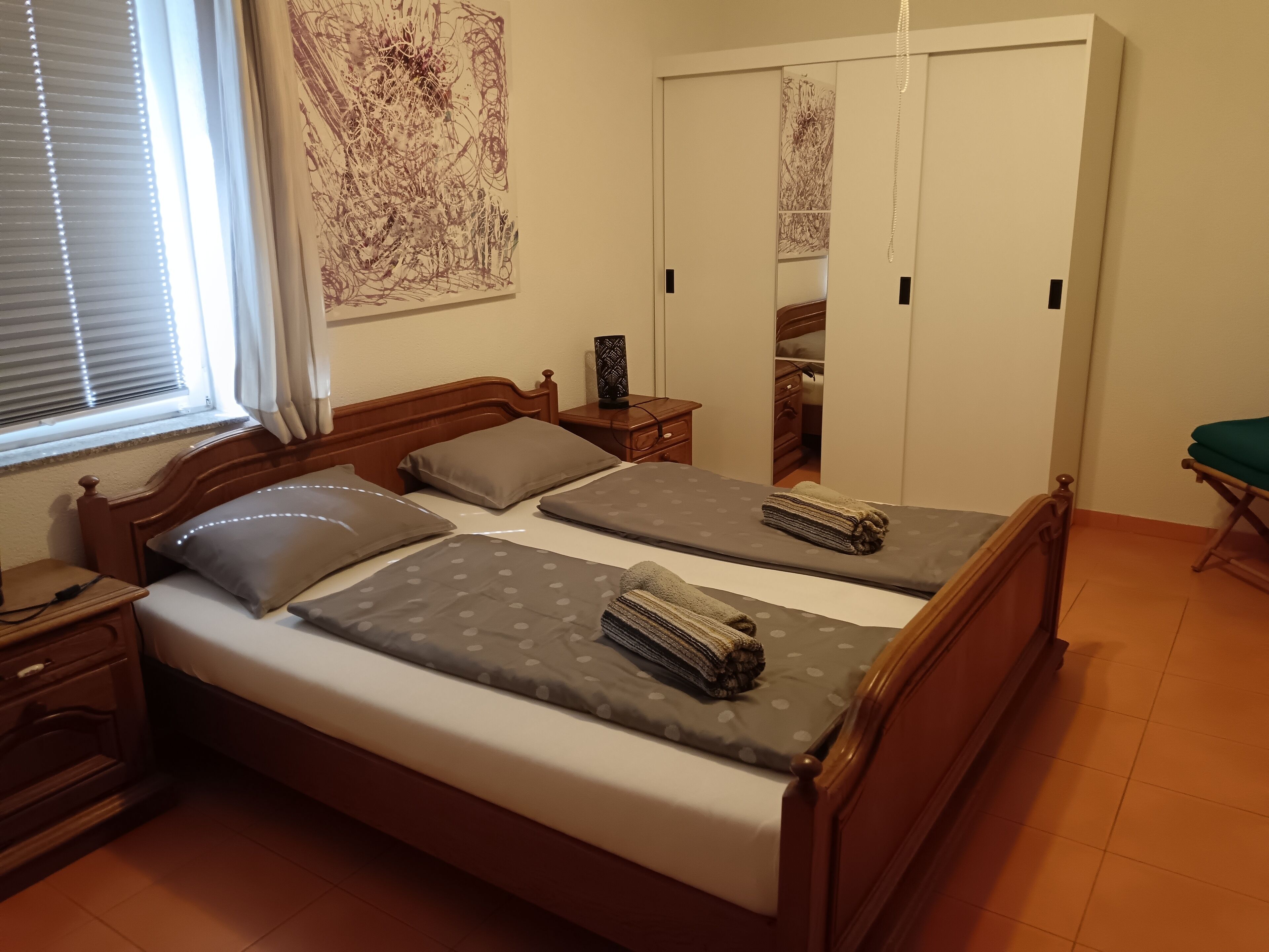 Un dormitorio con una cama de madera, dos almohadas, una mesita de noche con lámpara, un espejo y una ventana con persianas.