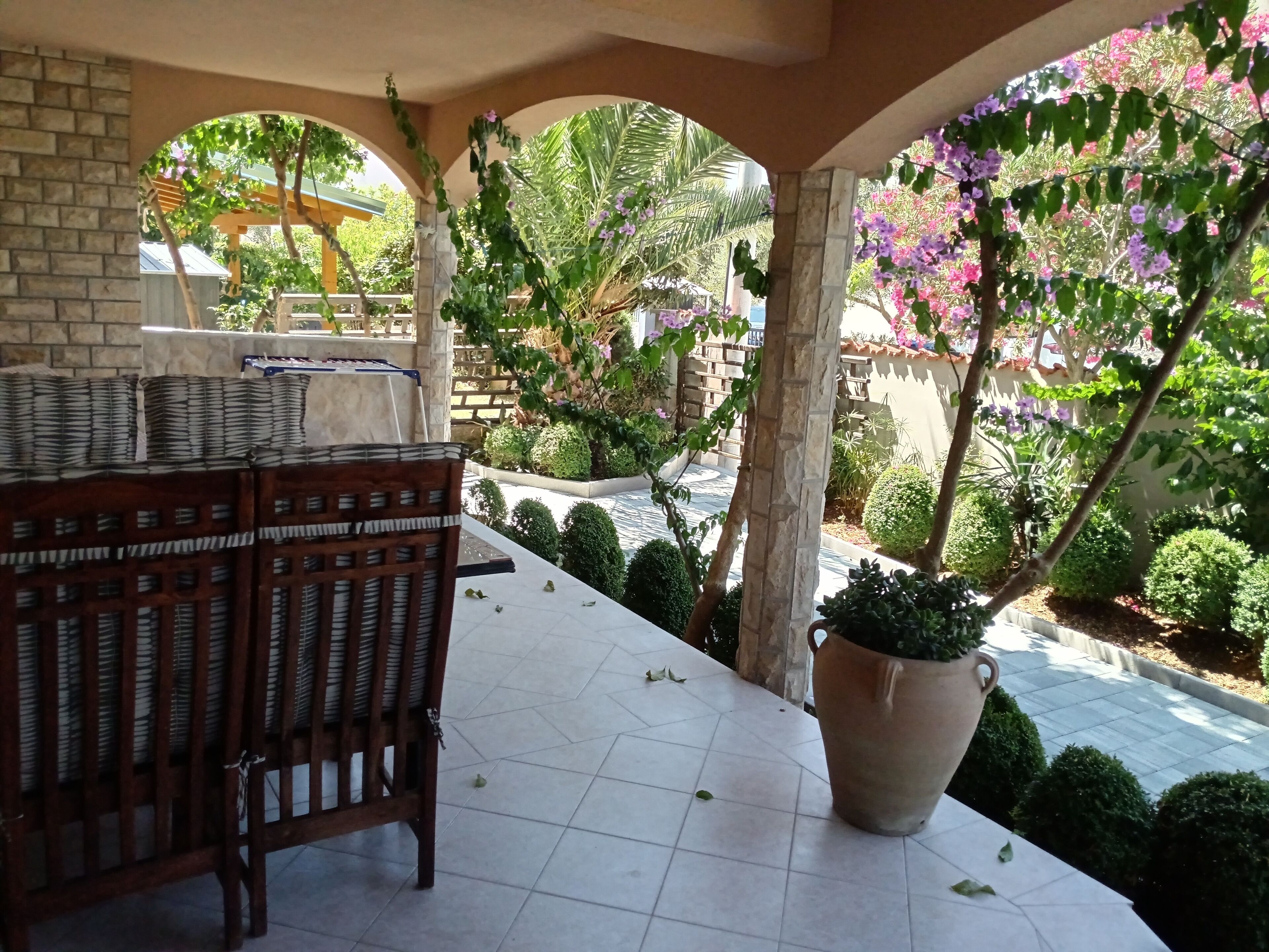 A covered patio area with a tiled floor, a wooden bench, and a large potted plant.