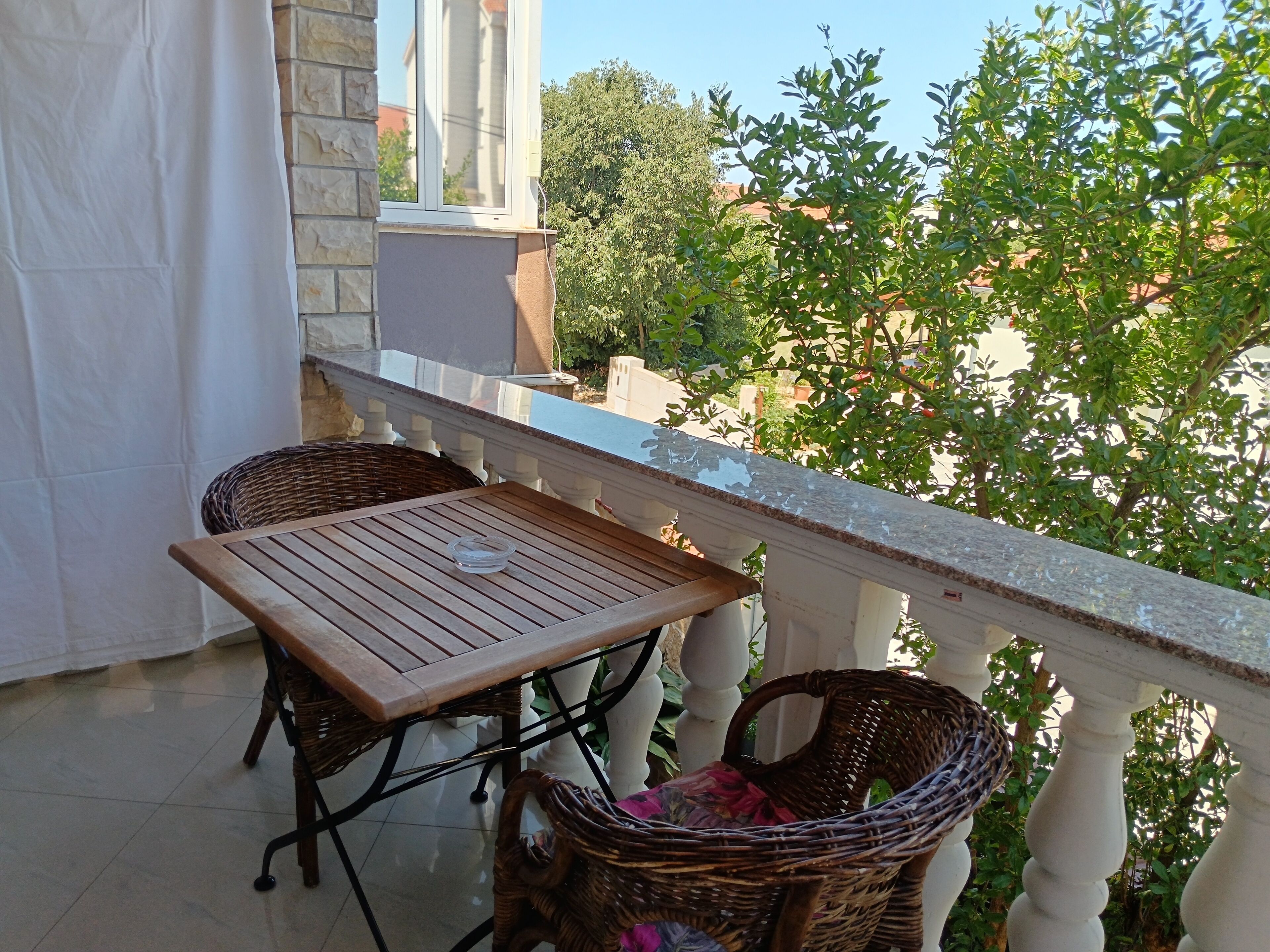 A balcony with a wooden table and wicker chairs, a white balustrade, and a view of greenery and buildings in the background.