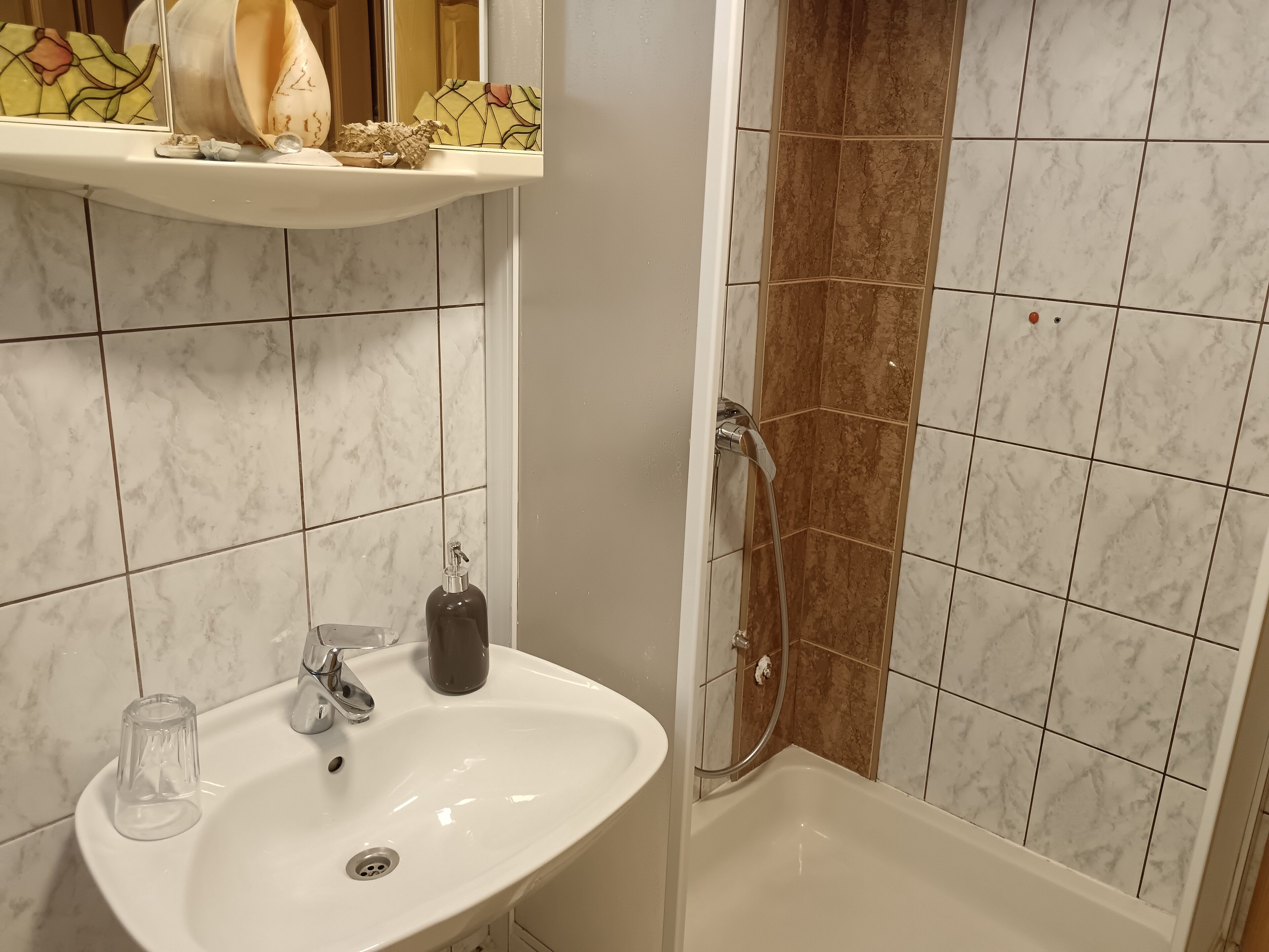 A bathroom with a white sink, a glass jar, a bottle, and a shower area with brown tiles.