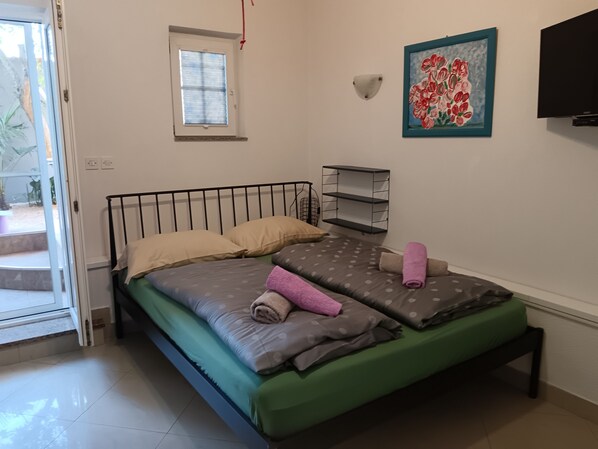 Habitación individual con una cama verde, varios almohadones, un ventanal pequeño, un estante en la pared y un cuadro de flores.