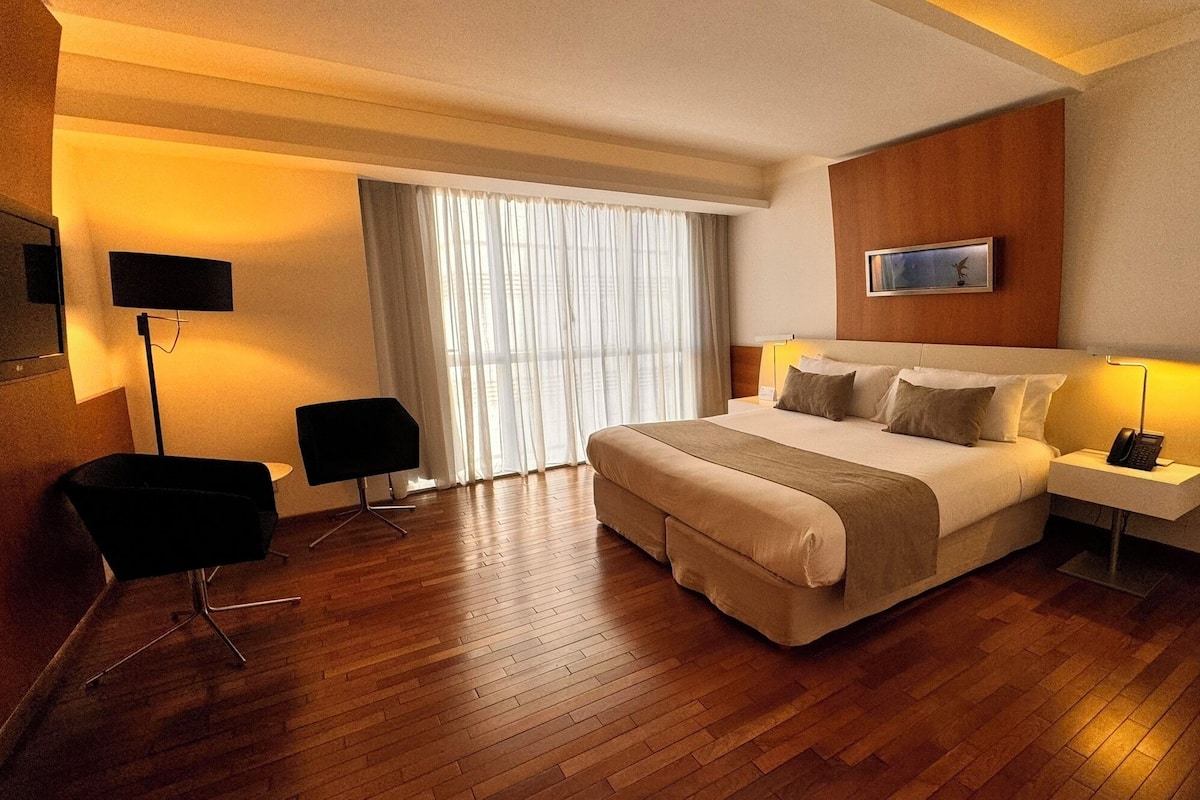 A hotel room with a large bed, a desk with a phone, a TV mounted on the wall, and a window with curtains.
