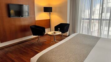 Ein Hotelzimmer mit einem großen Bett, zwei schwarzen Stühlen, einem kleinen Tisch mit Teller und Glas, einem flachbildfernseher an der Wand, einer Stehlampe und einem Fenster mit durchsichtigen Vorhängen.