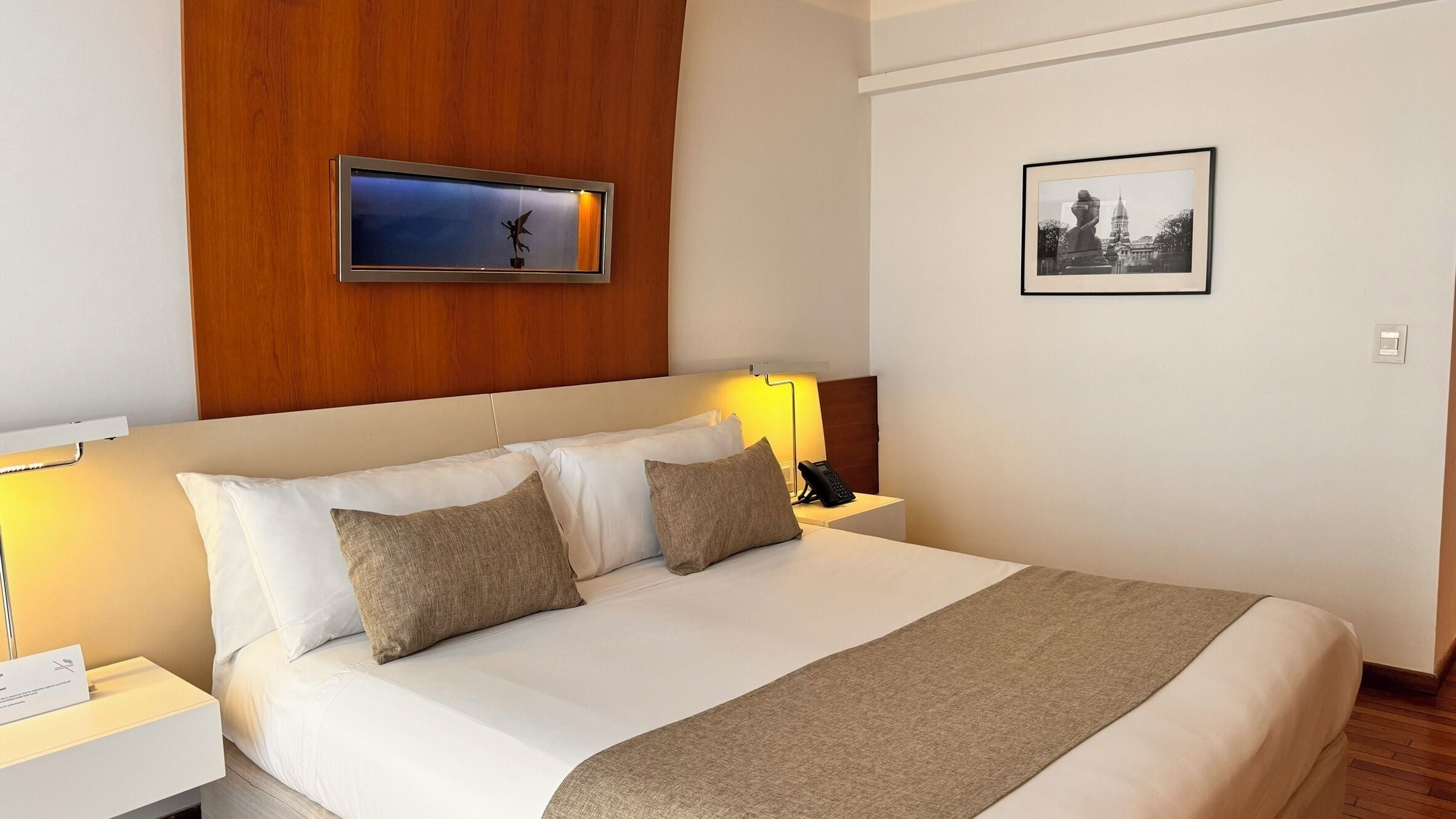 A hotel room with a large bed, two pillows, a bedside table with a lamp, a phone, a framed picture on the wall, and a wooden panel above the bed.