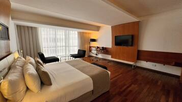 Habitación de hotel con una cama grande, un escritorio, una silla, un televisor y una ventana con cortinas.