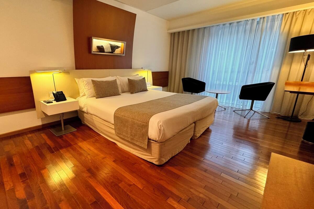 A hotel room with a large bed, bedside lamps, a small table, a chair, and a desk with a lamp.
