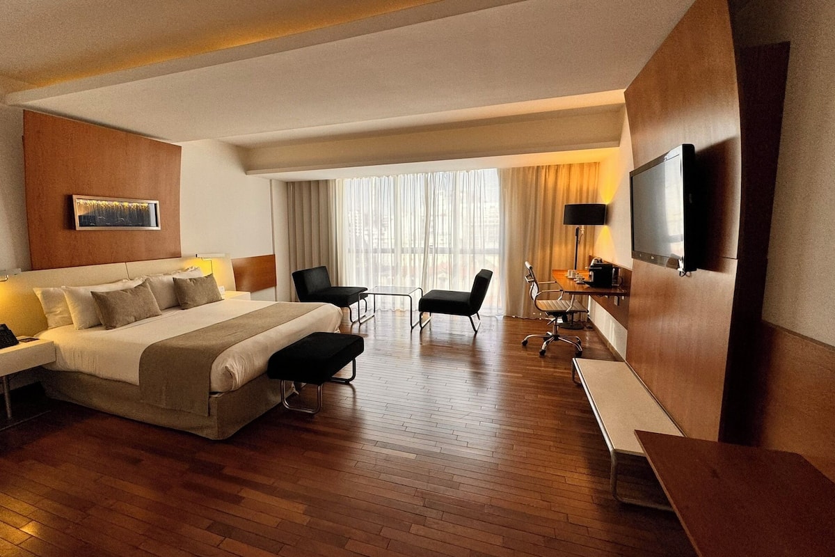 A hotel room with a large bed, bedside tables, a desk with a chair, a TV, and a window with curtains.
