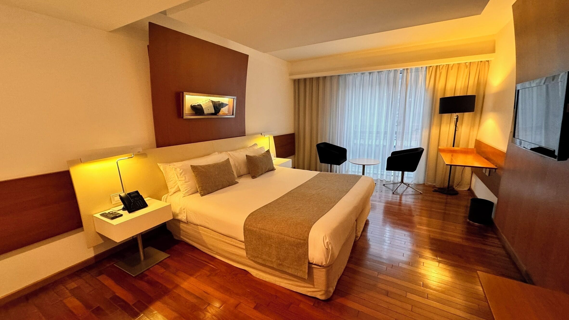 Una habitación de hotel con una cama grande, mesitas de noche con lámparas, un escritorio con una silla y un televisor en la pared.