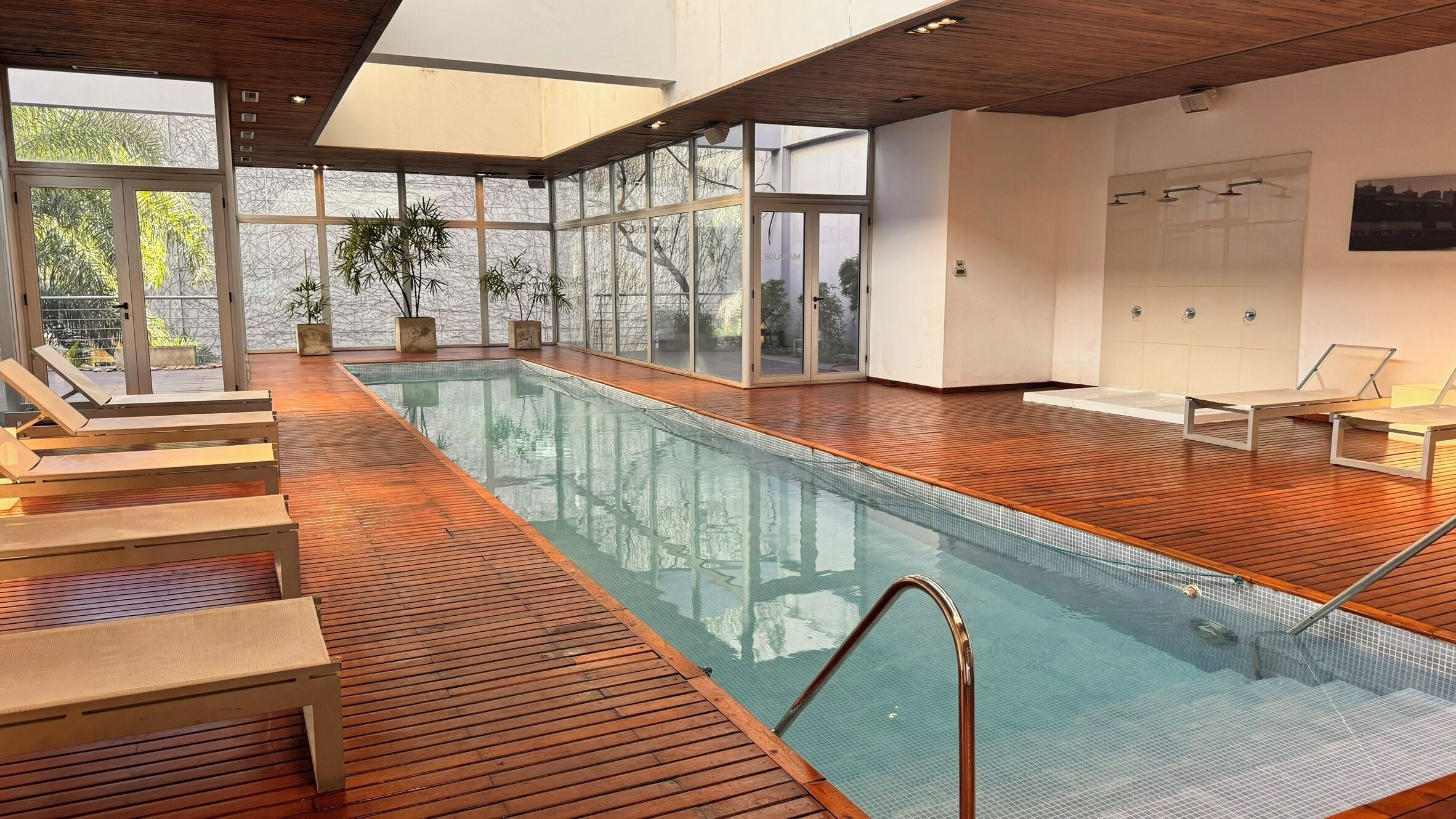 A modern indoor swimming pool with wooden flooring, glass walls, and lounge chairs.
