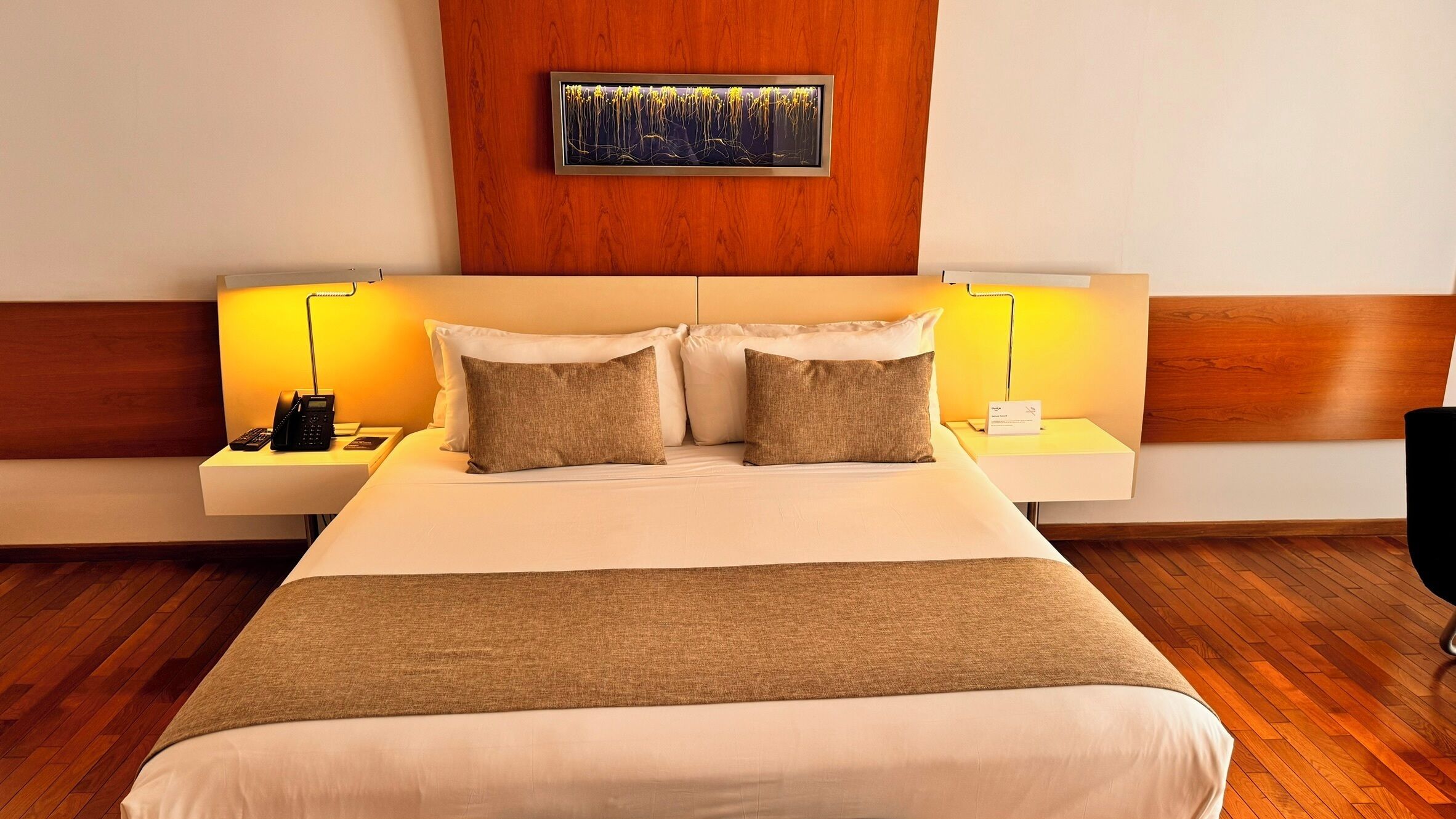 Une chambre d’hôtel avec un grand lit, deux tables de chevet avec des lampes, une tête de lit en bois et un tableau encadré au mur.