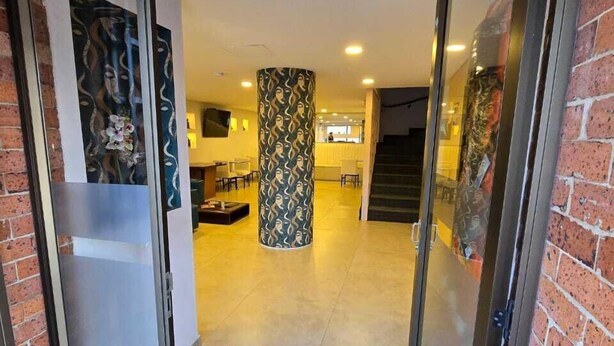 Un couloir avec une porte en verre donnant sur un espace d’accueil, un pilier orné de motifs et un escalier.